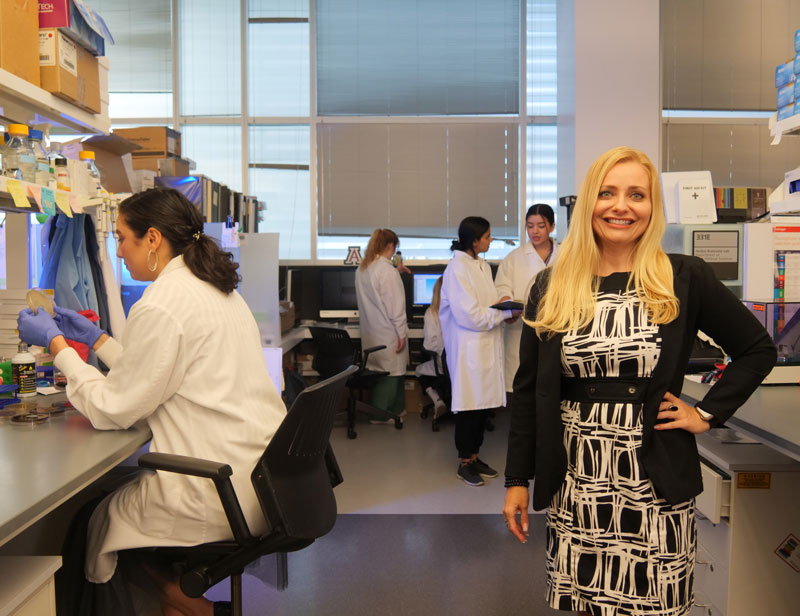 Woman with blonde hair and a dark and white dress stands with a hand on her hip while other women work in the lab behind her