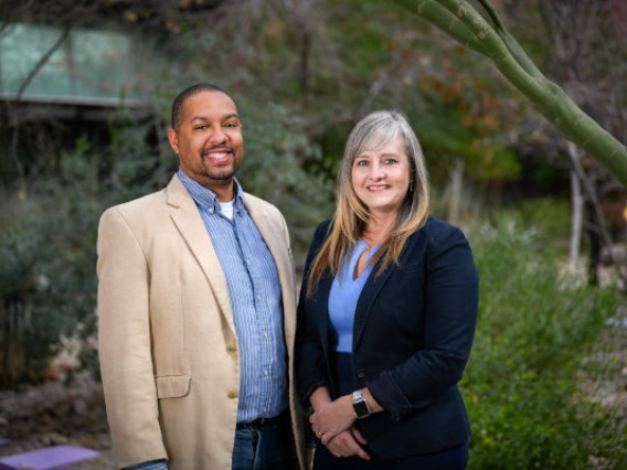 Black man in tan suit jacket and blue shirt next to a white woman with blue suit jacket and blue shirt. Both are smiling. Behind them are green trees. 