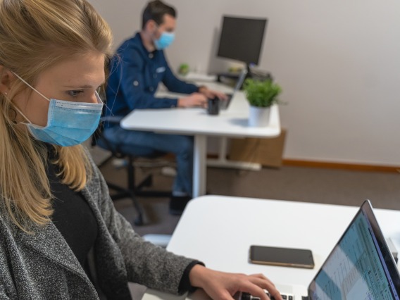Man and woman using laptops, sitting at separate desks wearing blue surgical masks