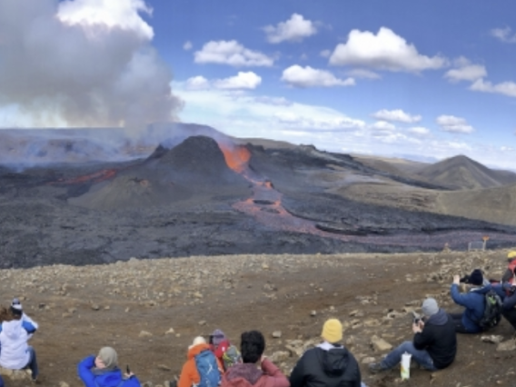 A volcano erupts in the background as researchers and onlookers watch in the foreground