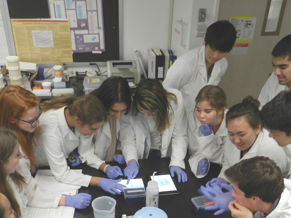 a large gropu of young people in lab coats and latex gloves gather around a tablet