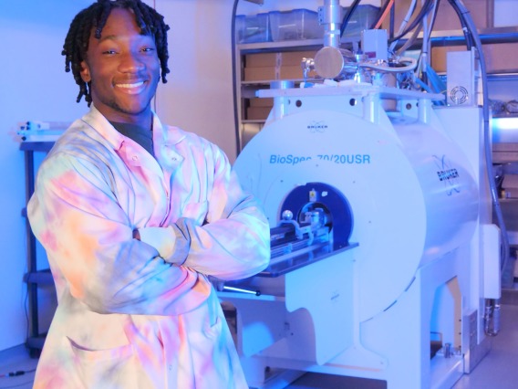Young black man wearing a tie dye lab coat crosses his arms in front of him. In the background is an MRI machine.