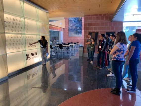 Woman stands in BIO5 Institute lobby wall and talks about membership at BIO5 with visiting students