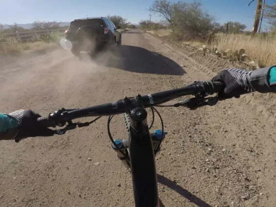 First-person view from a cyclist riding on a dusty road behind a moving vehicle, with handlebars and the bike’s front wheel visible in the forefront.