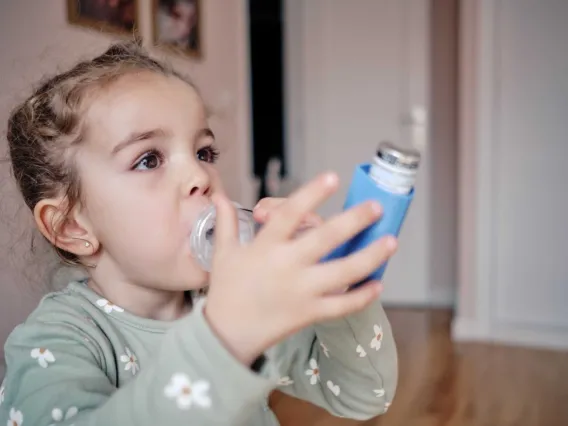 A young child drinks water from a blue insulated bottle. The child has pigtails and is wearing a light-colored shirt with flower patterns. The background shows a softly focused home interior.