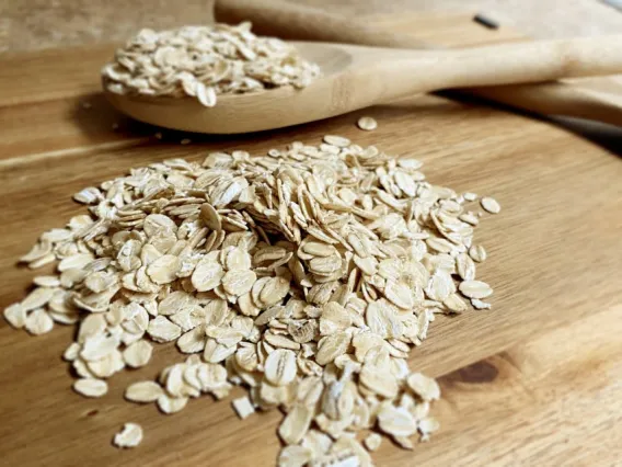 A close-up image of dry rolled oats scattered on a wooden surface, with a wooden spoon also containing oats resting on the surface. The focus is on the texture and natural appearance of the oats.