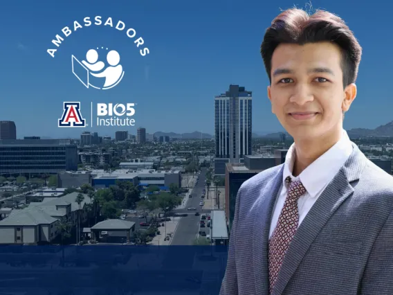 Professional portrait of an individual with short hair, wearing a suit, standing in front of a cityscape with the 'Ambassadors' logo, the University of Arizona (UA) emblem, and the BIO5 Institute logo visible in the background.