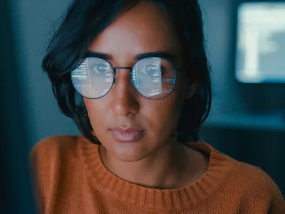 Person wearing glasses with computer screen reflections visible on the lenses, looking intently forward in a dimly-lit room.