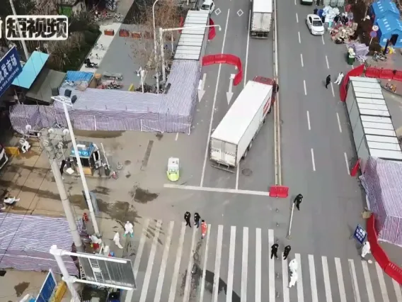 Aerial view of a busy street intersection with pedestrians crossing, surrounded by blue-roofed market stalls. Signage in Chinese characters is visible on nearby buildings.