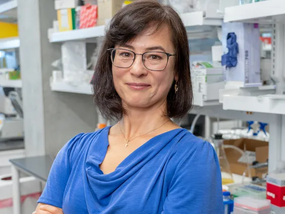 A scientist smiling at the camera, standing in a laboratory filled with equipment and supplies. The person is wearing a blue top and glasses.