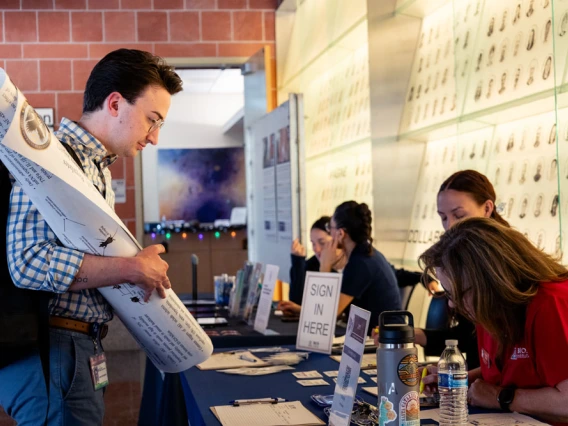 Person holding a poster interacts with another person seated at a booth labeled "Sign in Here" at a professional event.
