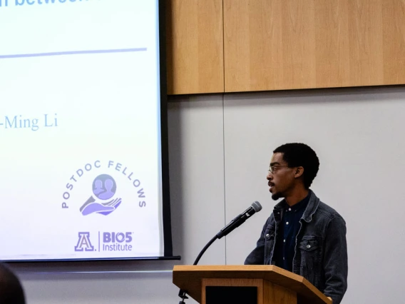 A person presenting at a lecture, standing next to a projector screen with text and the logo of the Postdoc Fellows at the BIO5 Institute