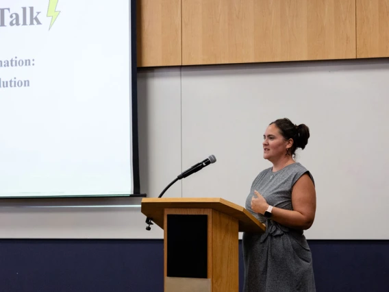 A person presenting at a lectern with a slideshow projected on a screen behind them, in a conference room setting.