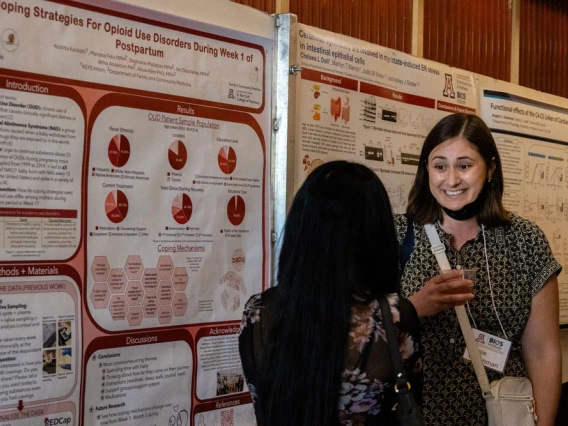 Two individuals at a scientific conference discuss in front of posters, which includes various graphs and text sections like Introduction, Results, and Methods & Materials.