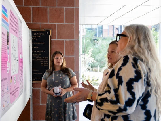 Three people discussing a presentation next to a poster board in a university building. One person is gesturing towards the poster while the other two listen attentively.
