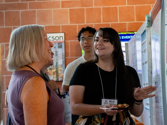Two individuals engaging in a conversation at a poster presentation session in a brick-walled, indoor setting. One person is gesturing while the other is listening attentively. All are wearing conference badges.