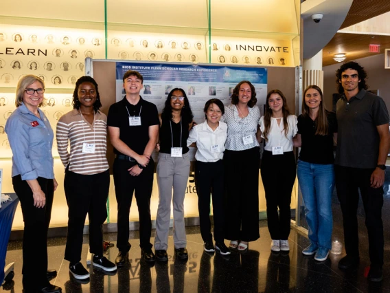 Group of eight individuals standing in front of a display with headshots, smiling and posing for a photo.
