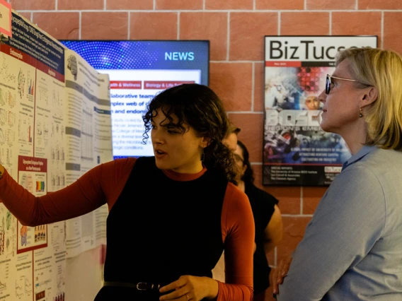Two individuals examining and discussing a research poster at a conference. One person is pointing at the poster, which shows various charts and text, while the other listens attentively. 