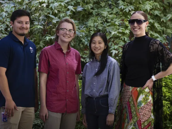 Four people smiling and standing together outdoors, with greenery in the background. From left to right, one person is wearing a blue polo shirt, the next is in a red button-down, followed by a person in a light blue shirt, and another in a black top with lace sleeves.