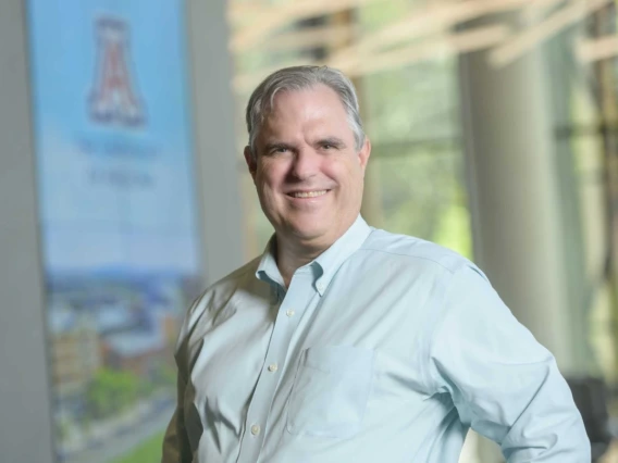 Individual smiling in front of a banner displaying 'The University of Texas at Arlington,' set against a light-filled, blurred background of a building interior.