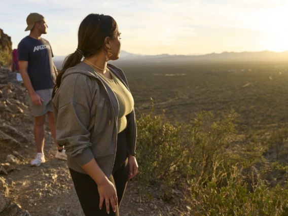 Two people standing on a rocky trail, overlooking a vast desert landscape at sunset. One individual is wearing a hat