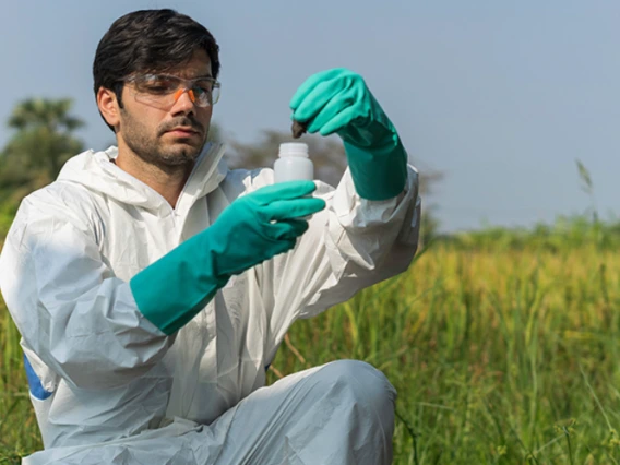 A scientist in a white lab coat and gloves examining a sample in a flask outdoors in a grassy field.