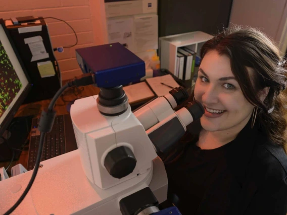 Person smiling at camera while working with a microscope in a laboratory.