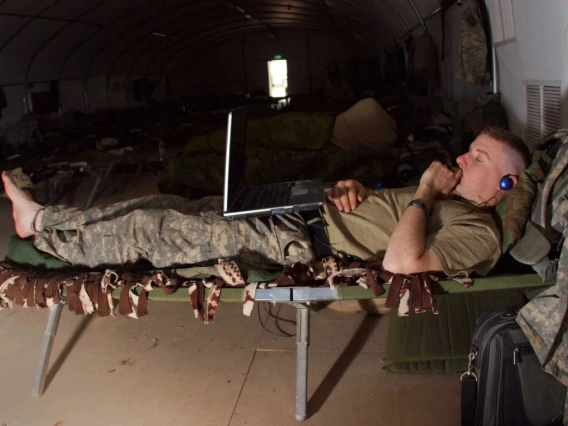 A person in military uniform is reclining on a cot in a tent, using a laptop and talking on a phone. The environment appears to be a temporary military camp setup.