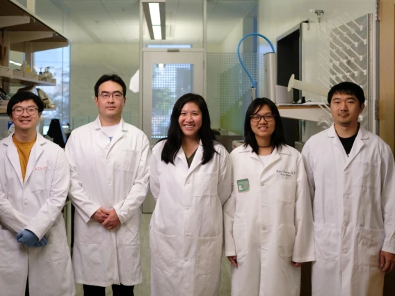 Five laboratory technicians wearing white lab coats are standing in a research lab. Each individual is smiling and facing the camera.