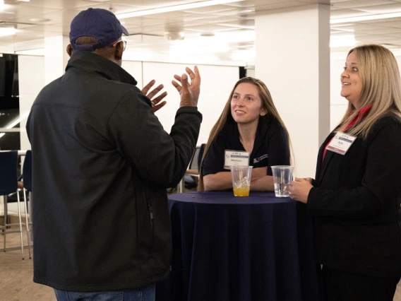 Three individuals engaging in conversation at a networking event, with two listeners wearing name tags and standing behind a table with a drink.