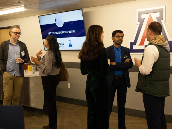 Five individuals are engaged in conversation at a networking event at the University of Arizona, as indicated by the logo on the screen displaying "Let the Networking Begin." 