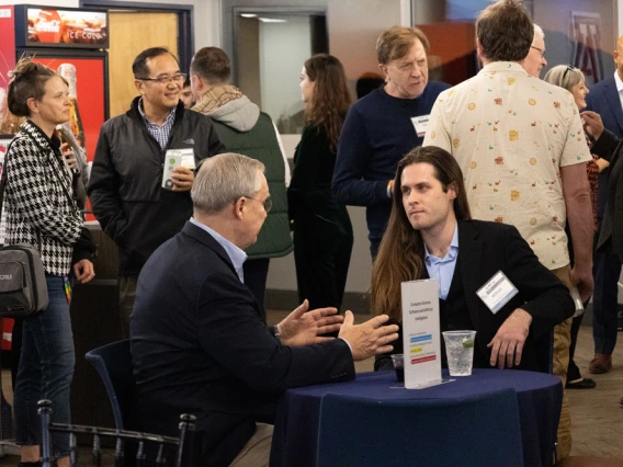 A networking event at a conference with attendees engaging in discussions. Two sit at a table and other individuals converse in the background near refreshment stands.