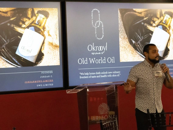 Person presenting at a podium with two projected screens behind them displaying advertisements for "Okroyl Old World Oil." The screens show images of an oil bottle.