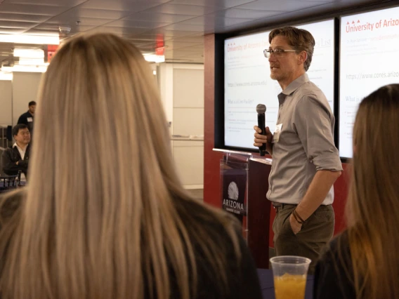 A person presenting at a University of Arizona event, standing with a microphone in front of a projected screen, addressing an audience.