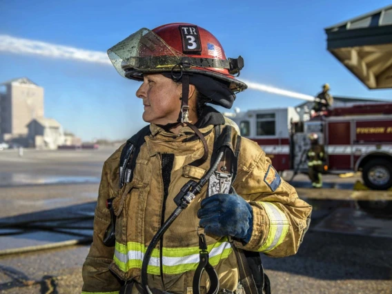 A firefighter in full gear stands in front of a fire truck labeled "Fresno Fire," with industrial buildings in the background. The firefighter is holding a helmet under the left arm and a radio in the gloved right hand.