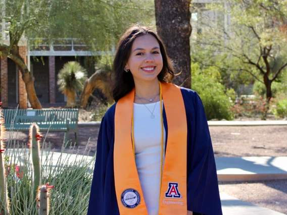 A person wearing a University of Arizona engineering graduation stole stands smiling outdoors in a sunny campus setting.