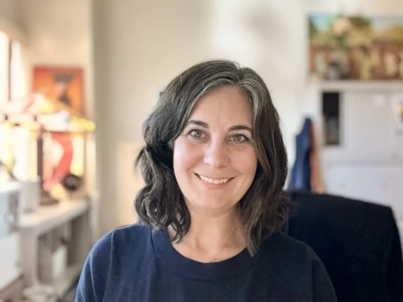 Portrait of a person smiling, with shoulder-length curly hair, wearing a blue top, in a well-lit room featuring art supplies and paintings in the background.