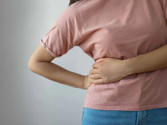 Person in a pink shirt holding their lower back, possibly indicating back pain, against a plain grey background.