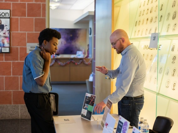 Two individuals discussing over a desk in an office environment, with posters on the wall including one reading "Tucson.