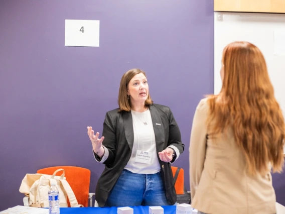 Person at a networking event, speaking animatedly to another attendee at a table with papers and cups, in a room with numbered stations on the walls.