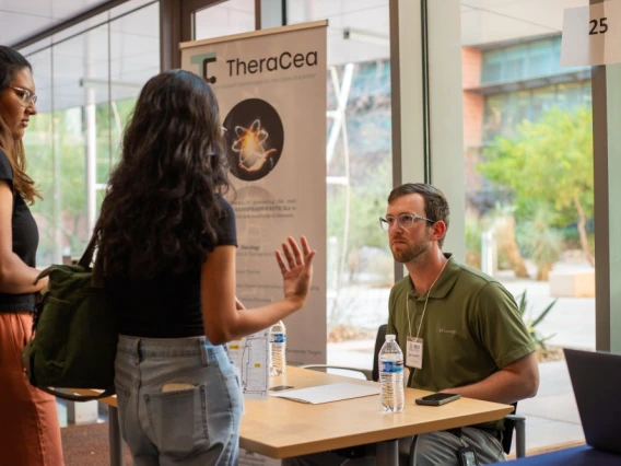 Three individuals discussing at a TheraCea exhibit booth inside a building with large windows and modern architecture.