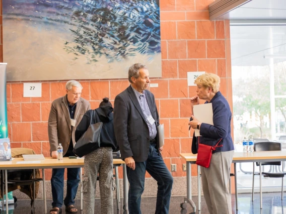 Three individuals are engaged in a conversation at a conference beside numbered booths, under a large abstract painting.