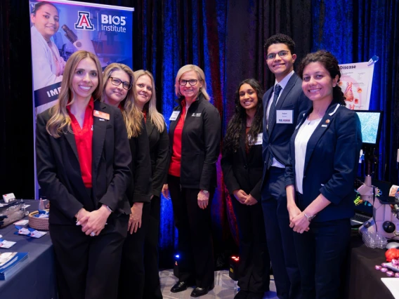 Group of seven individuals standing with confident smiles at a University of Arizona BIOS Institute recruitment booth, where banners and promotional materials are visible in the background.