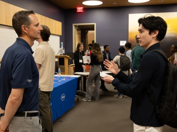 Two individuals are engaged in a conversation at a career fair with a Roche banner visible in the background. Other attendees are interacting and networking around them in a room with vibrant decor.