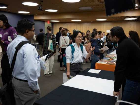 Attendees engaging with a presenter at a conference booth inside a room with wood-paneled walls and informational materials displayed on the table.