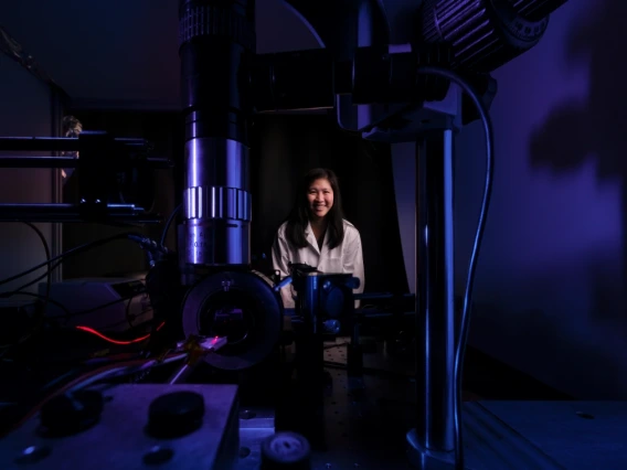 A researcher smiling in a dark laboratory setting, surrounded by advanced optical equipment including lasers and lenses.