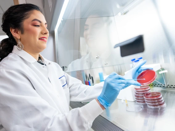 Scientist in a lab coat examining a petri dish in a laboratory, wearing blue gloves and smiling.