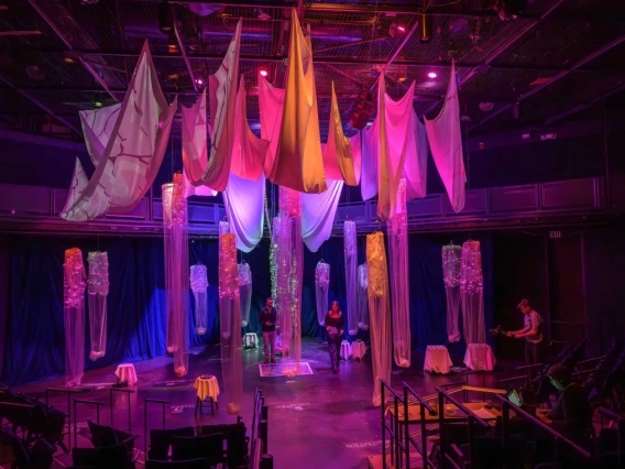 Interior of a theater with colorful hanging fabrics and illuminated set pieces, with two individuals seated at opposite ends of the stage.