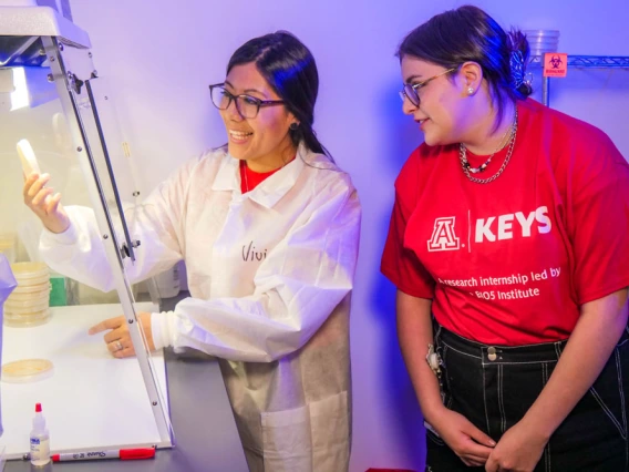 Person in white lab coat holds up petri dish underneath a hood while a person in a red KEYS t-shirt looks on