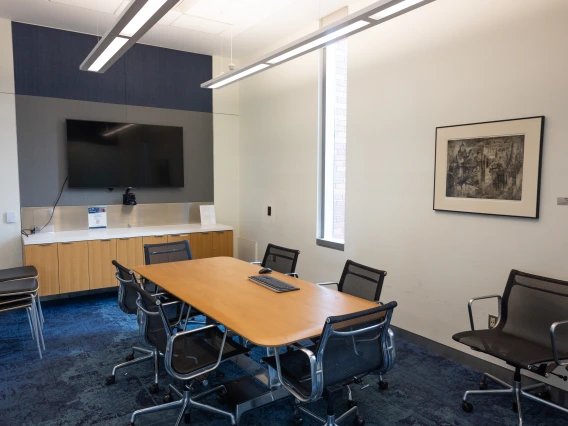 Conference room with a large wooden table, surrounded by chairs, a presentation monitor on one wall, and a framed cityscape artwork on another wall.
